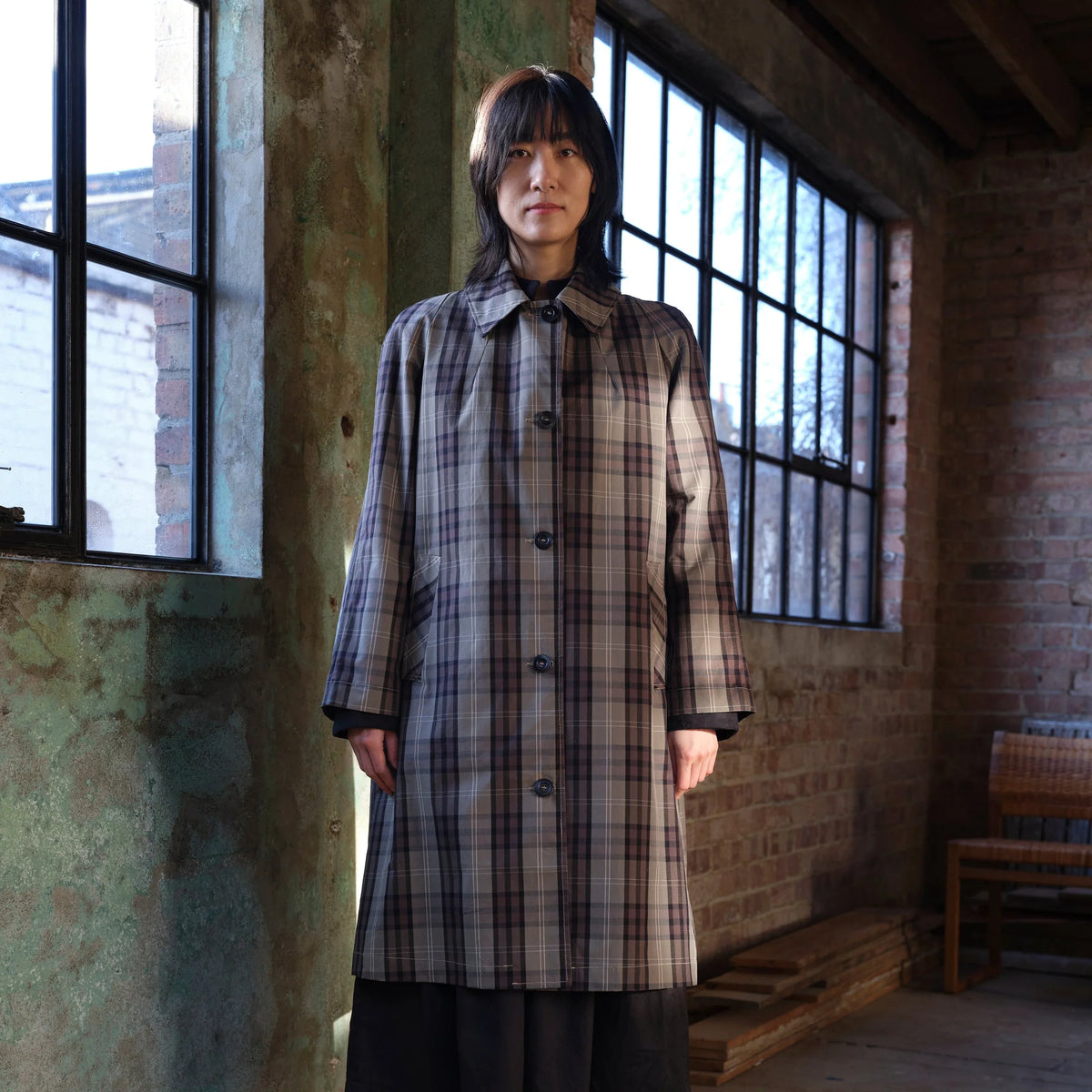 A person stands indoors wearing a long plaid coat in muted beige, gray, and brown tones, with dark bottoms. The setting is a rustic room with large black-framed windows, exposed brick walls, and a wooden bench.