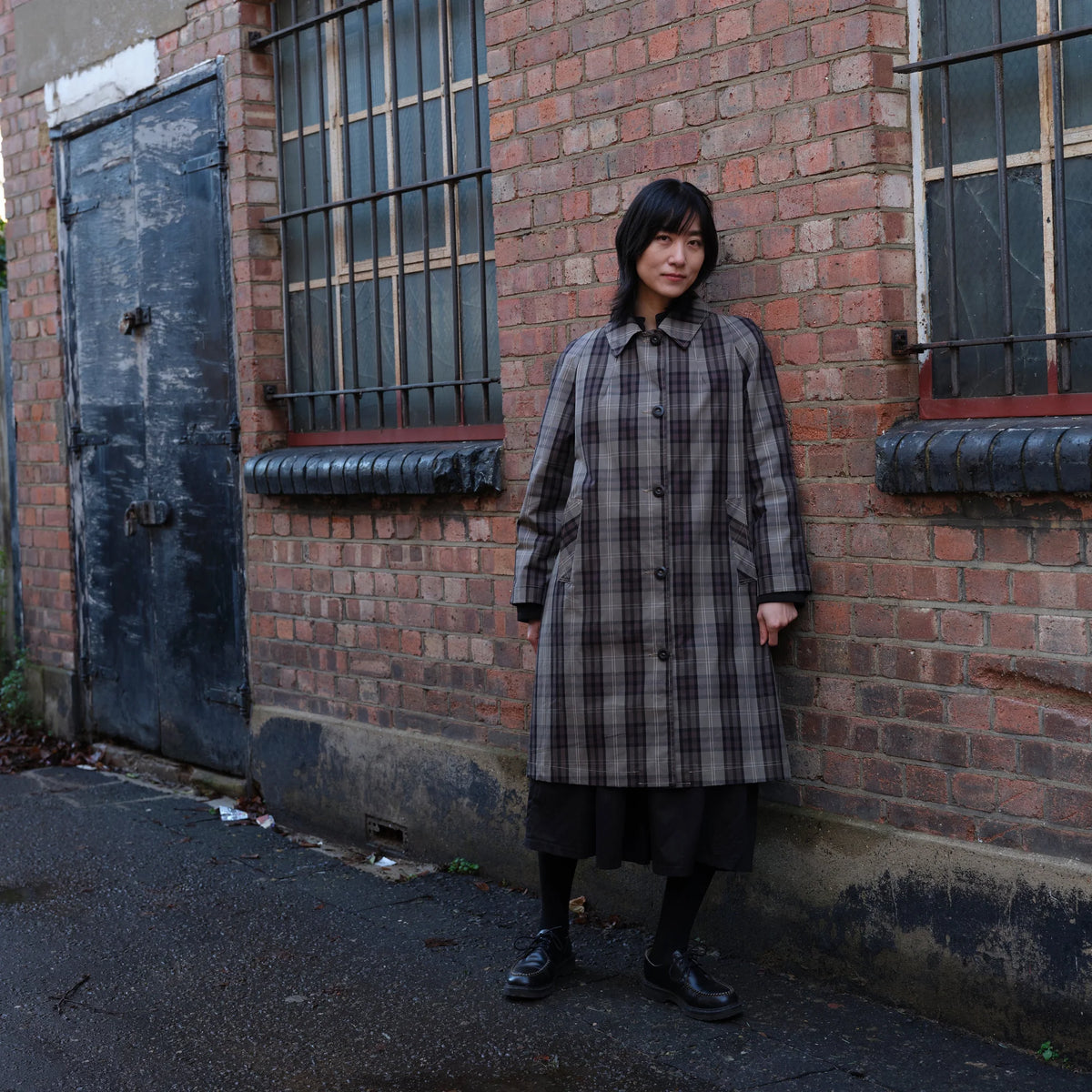A woman stands against a red brick wall wearing a long gray and black plaid coat over a dark skirt, with black socks and shoes.
