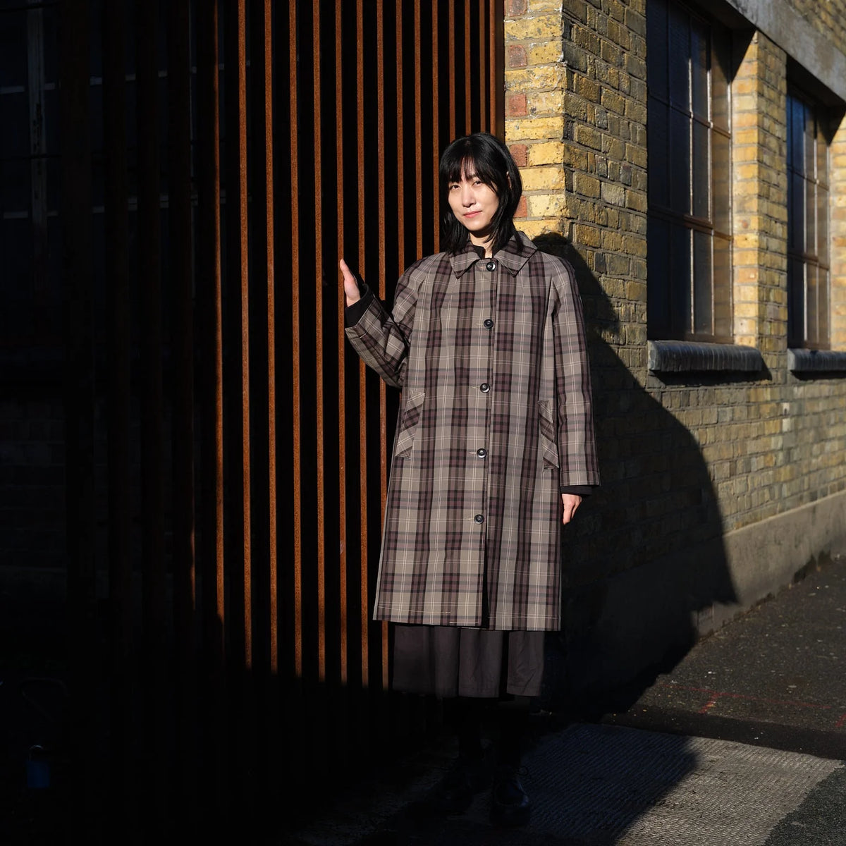 A woman stands outdoors against a brick building and dark slatted wall, wearing a long plaid coat over a dark dress and boots.