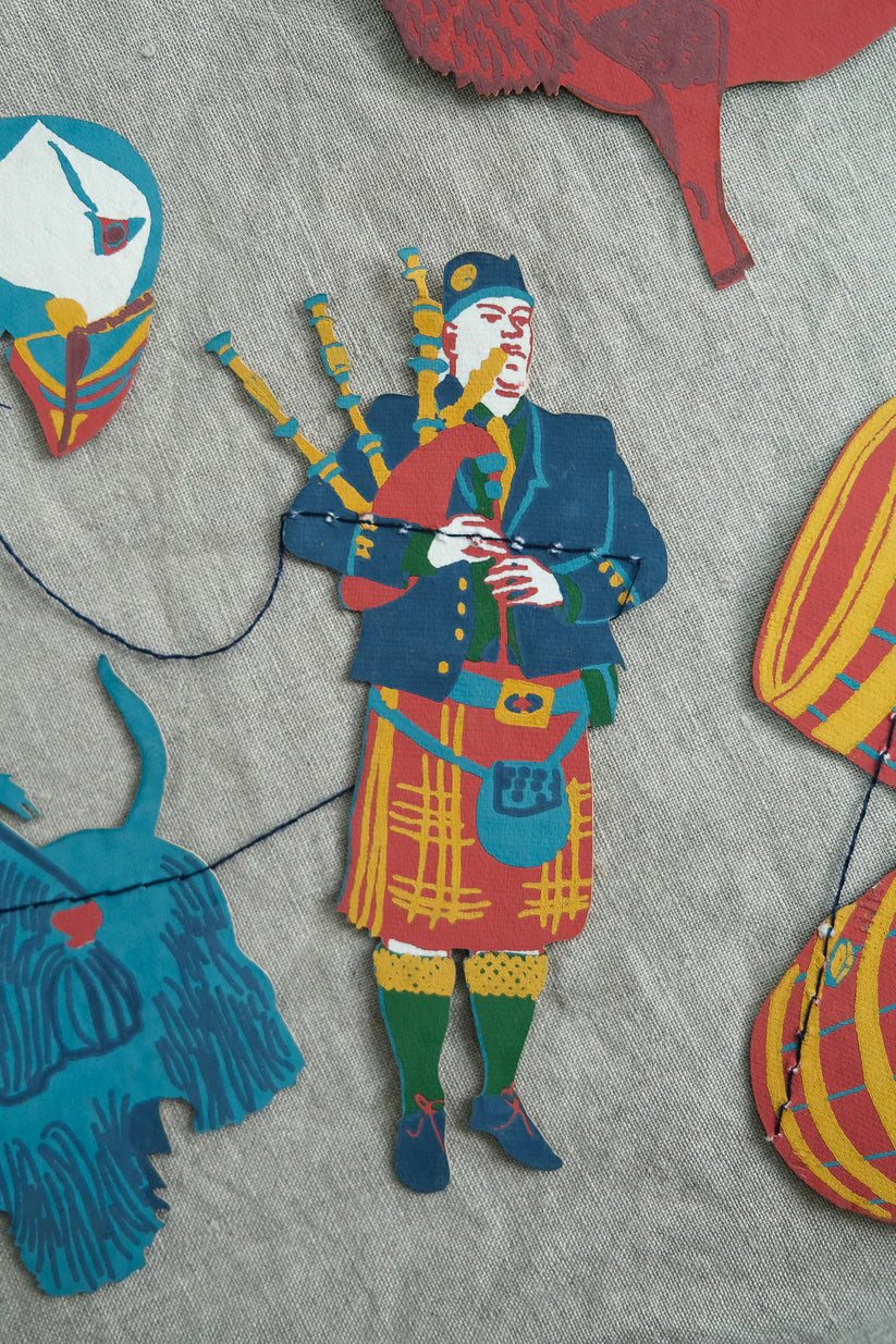 A colorful paper cutout of a bagpiper in a kilt, part of a garland featuring other Scottish-themed figures.