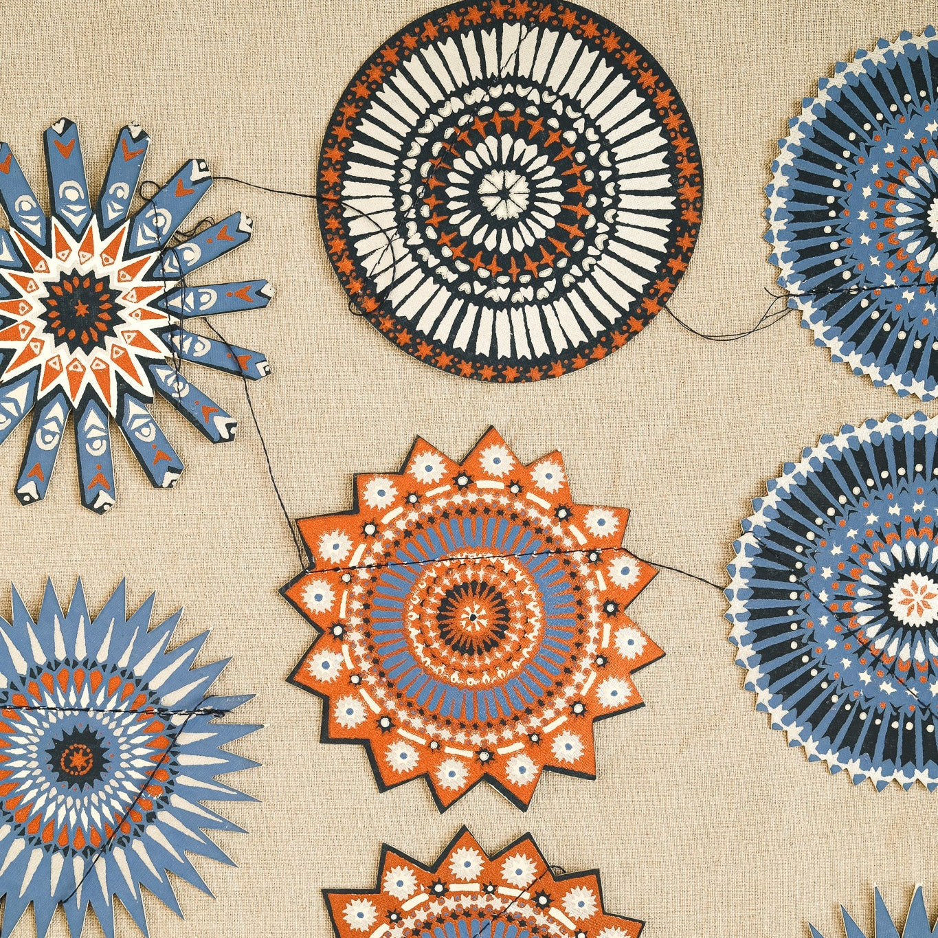 A decorative garland of circular and starburst paper ornaments with intricate mandala patterns in blue, orange, black, and white, strung along thin black cords against a beige fabric background.