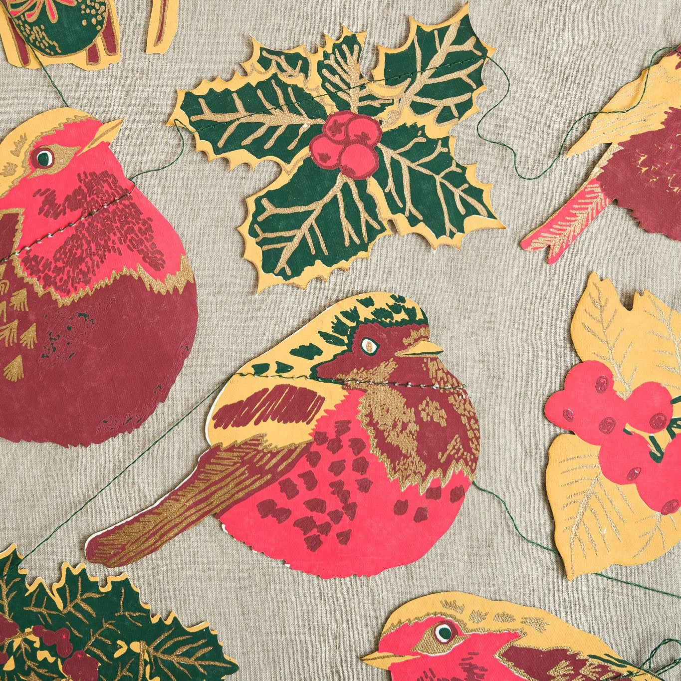 A close-up of a fabric or wallpaper pattern featuring vintage-style birds, holly leaves, and floral motifs in red, green, yellow, and brown on a light gray textured background.