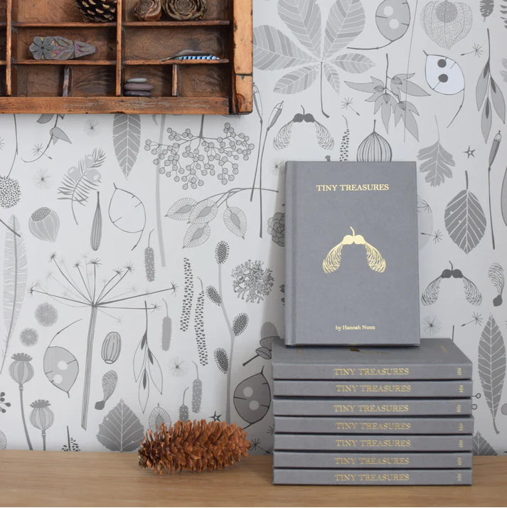 Stack of gray "Tiny Treasures" books by Hannah Nunn, featuring gold leaf detail, against a botanical wallpaper.