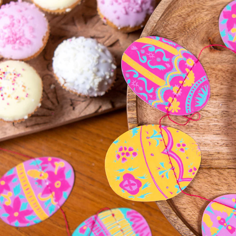 Colorful Easter egg-shaped decorations on a wooden surface with cupcakes in the background.