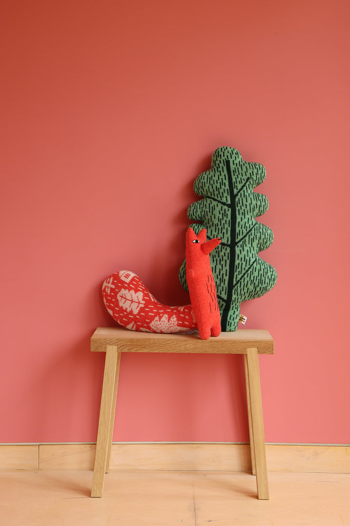 A red fox plush and a green tree cushion by Donna Wilson sit on a wooden table against a pink wall.