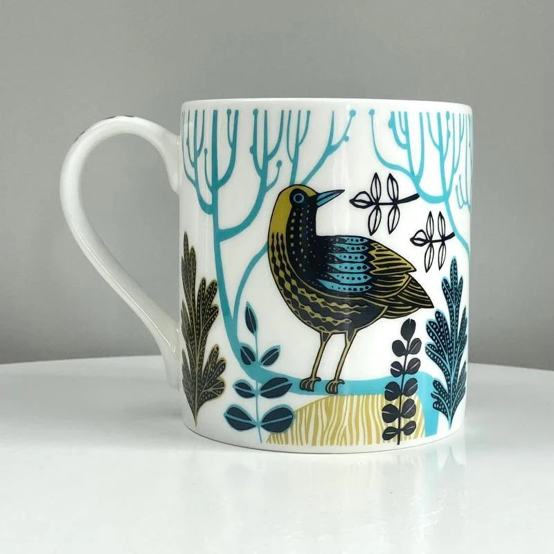 White mug featuring a stylized bird illustration in teal, black, and gold, surrounded by stylized foliage.