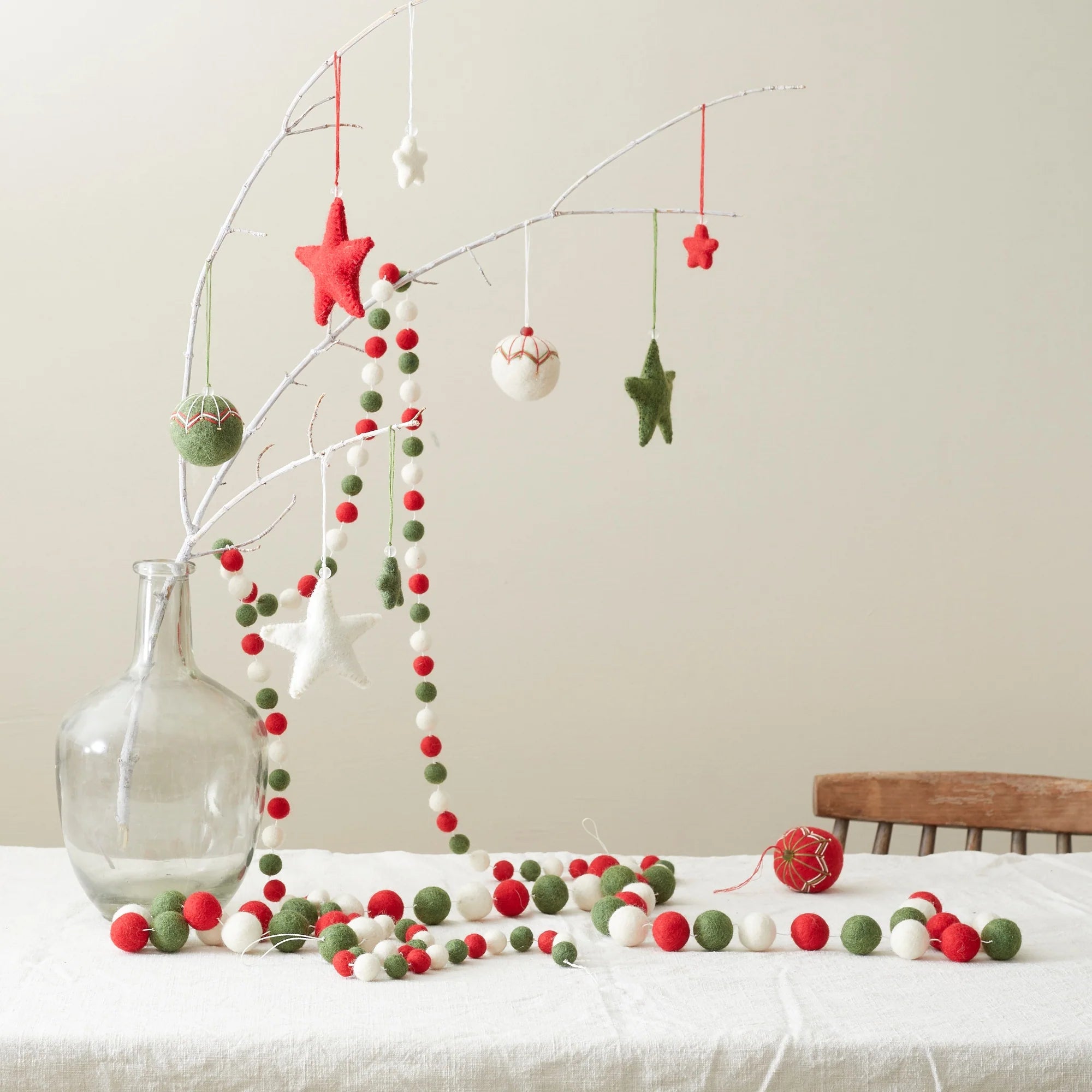 Red, green, and white felt star and ball ornaments hanging from a branch in a glass vase, with a matching felt ball garland.