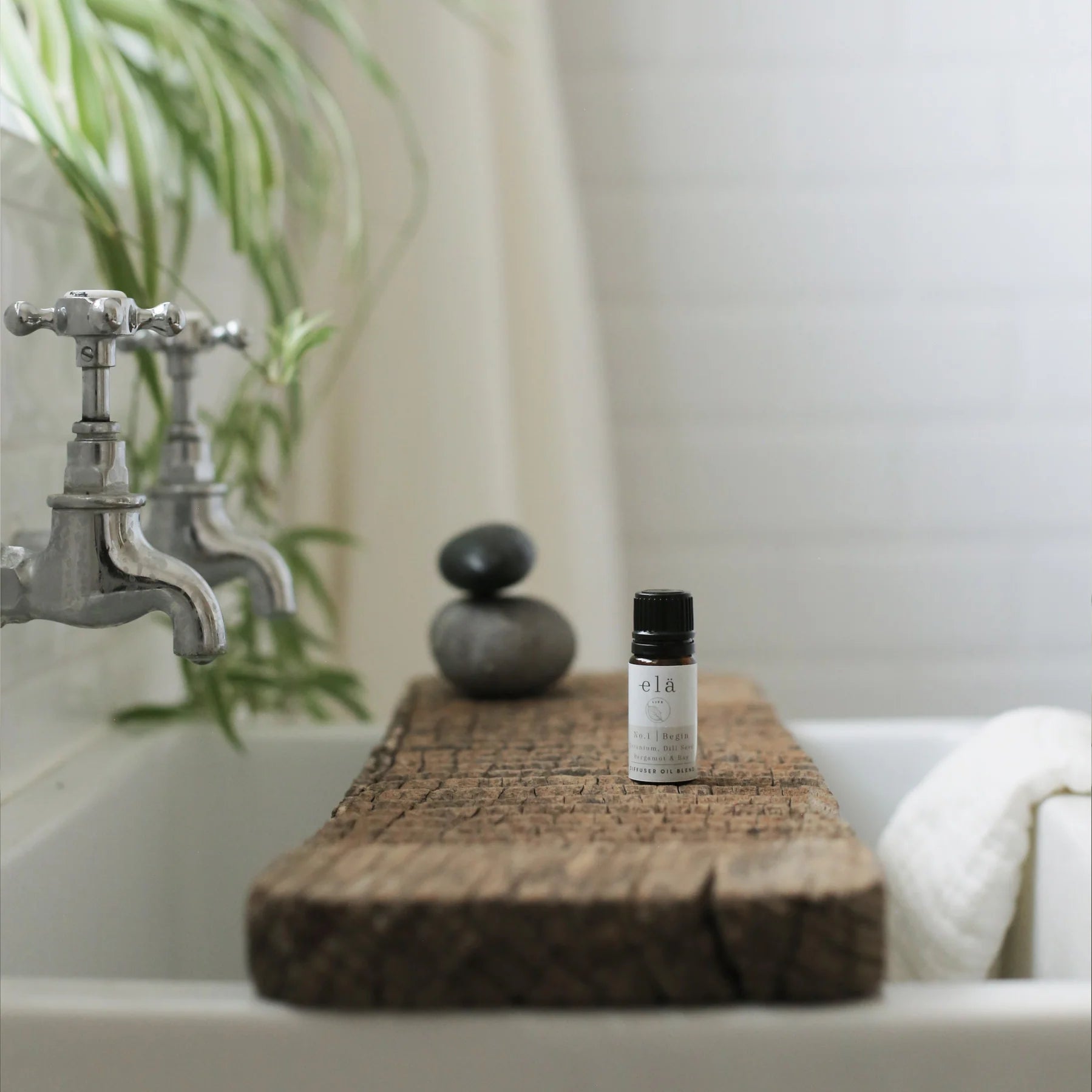 elá No.1 Begin diffuser oil blend in a dark glass bottle, resting on a rustic wooden board above a white sink.