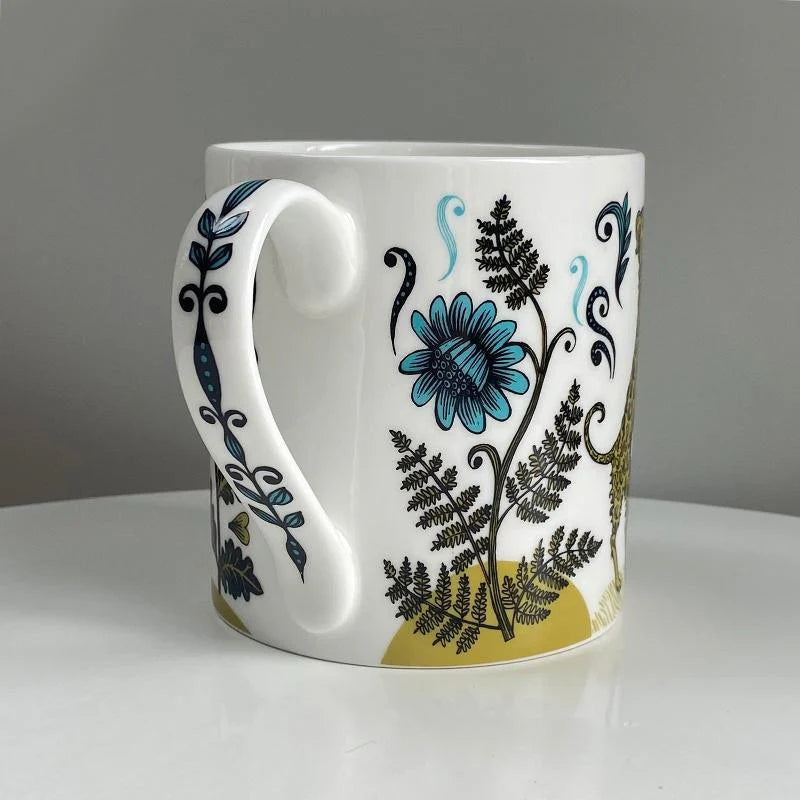 White mug with a large, curved handle, featuring a teal flower and fern design on a mustard yellow background.