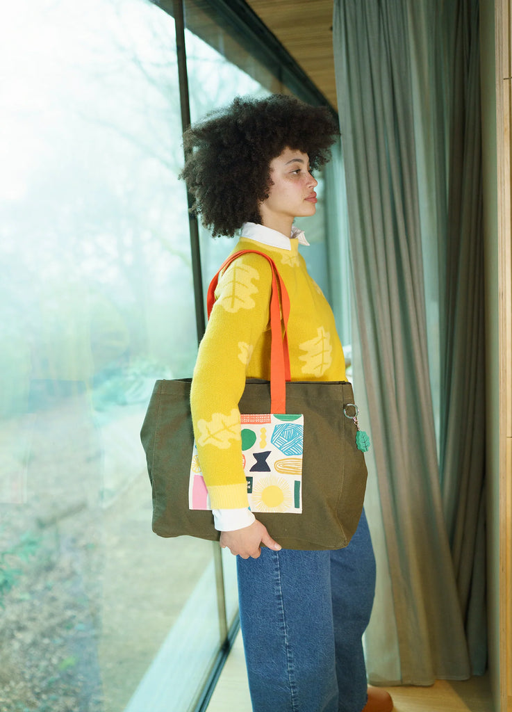 A woman with curly hair wears a yellow sweater by Donna Wilson, holding a green bag with colorful patterns and orange straps.