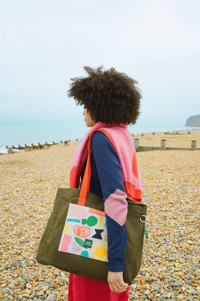 A woman in a pink and red sweater, blue top, and olive bag by Donna Wilson stands on a pebbly beach.