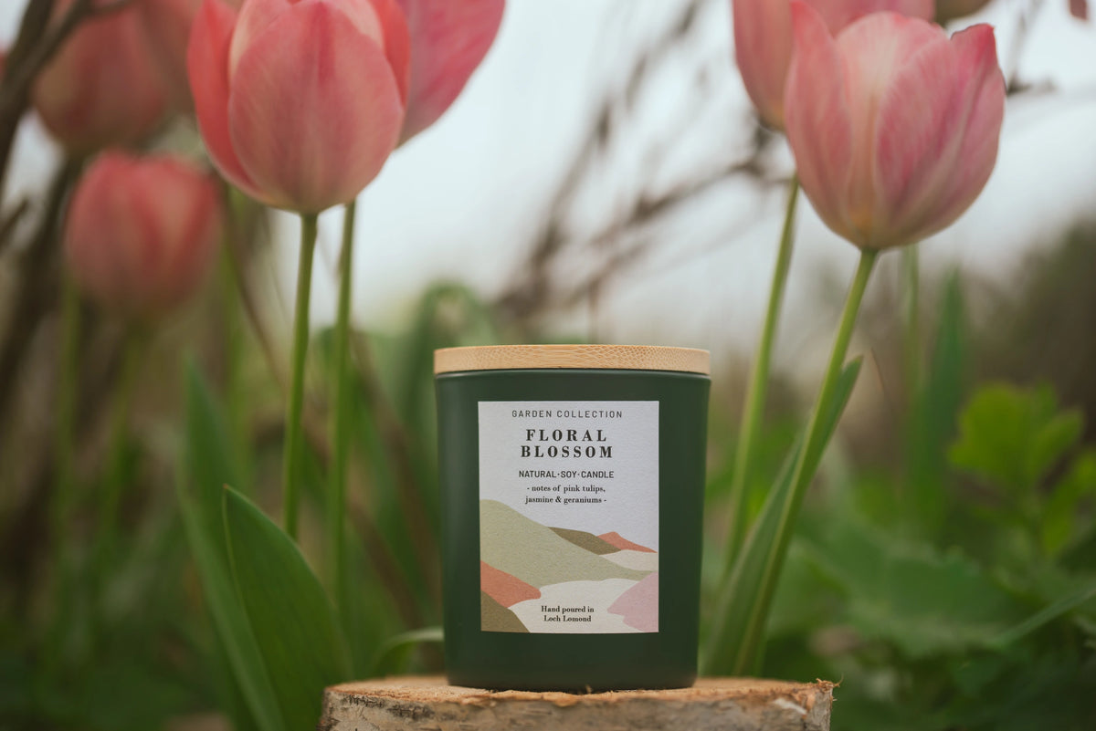 A dark green jar candle with a light wood lid sits on a small tree stump in a garden setting with pink tulips in the background. The front label reads 'GARDEN COLLECTION' and 'FLORAL BLOSSOM'.