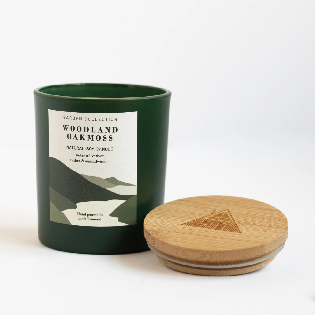A dark green soy candle jar with a white label reading 'Woodland Oak Moss' from the Garden Collection, shown next to its round wooden lid on a clean white background.
