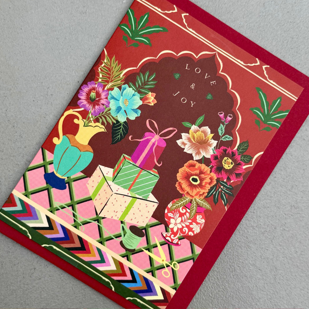 A square greeting card featuring a festive illustration with stacked gift boxes, colorful flowers in vases, and the words 'LOVE & JOY' on a red background with gold decorative borders.