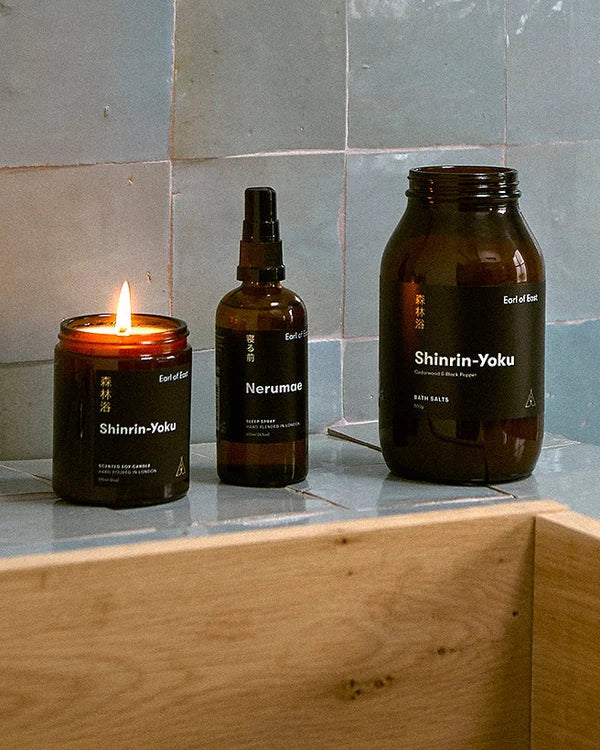 Three amber glass wellness products on a stone surface: a lit candle labeled Shinrin-Yoku, a spray bottle labeled Nerumae, and a larger bottle labeled Shinrin-Yoku, with a light wood box in the foreground and pale stone tiles behind.