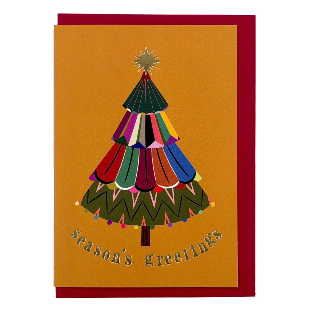 A Christmas greeting card with a mustard yellow front featuring a colorful stylized Christmas tree and the words 'season's greetings' in metallic script, shown with a dark red envelope behind it on a white background.