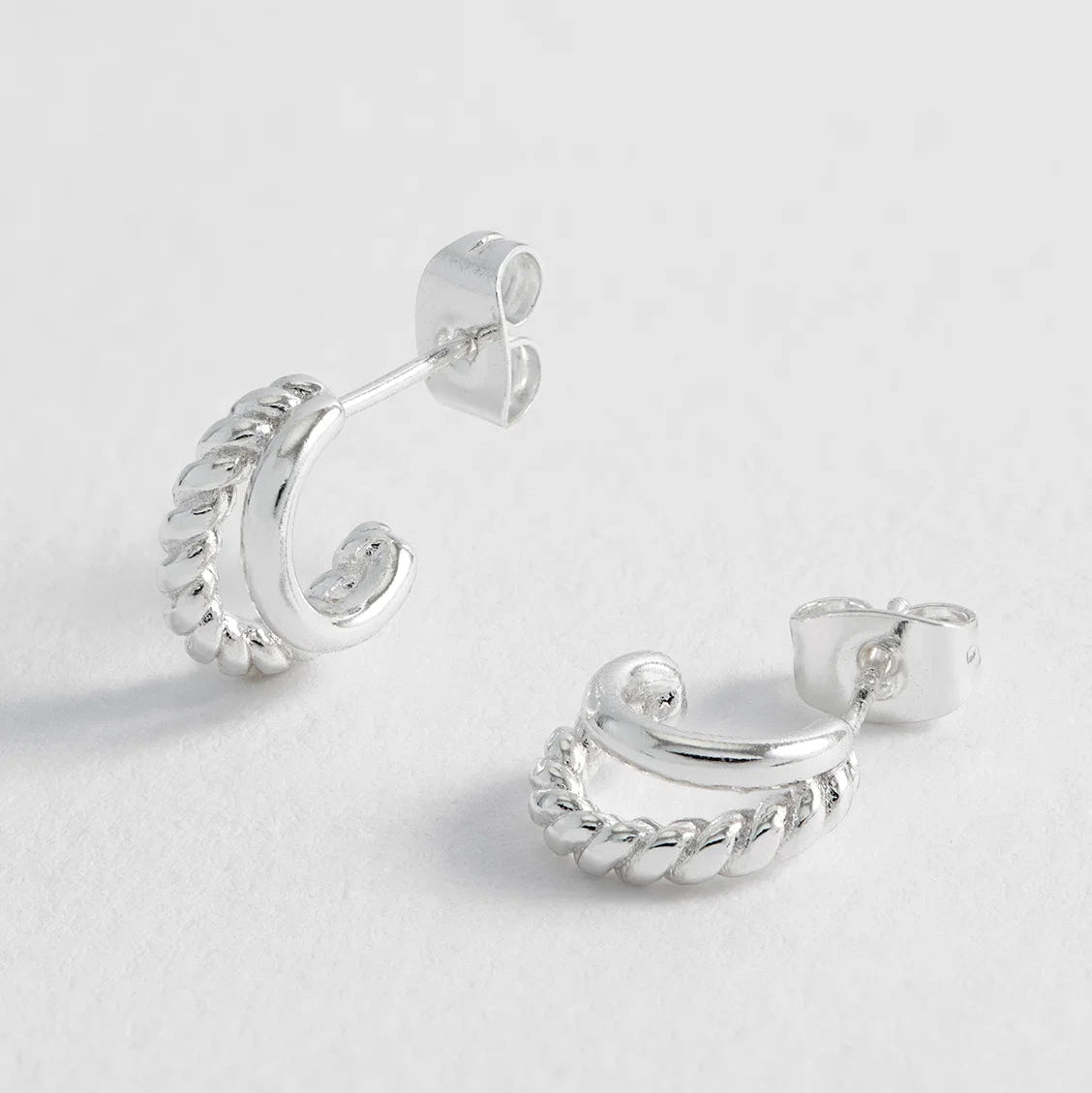 A pair of silver huggie hoop earrings with a textured twisted rope design, displayed on a clean, light gray background.