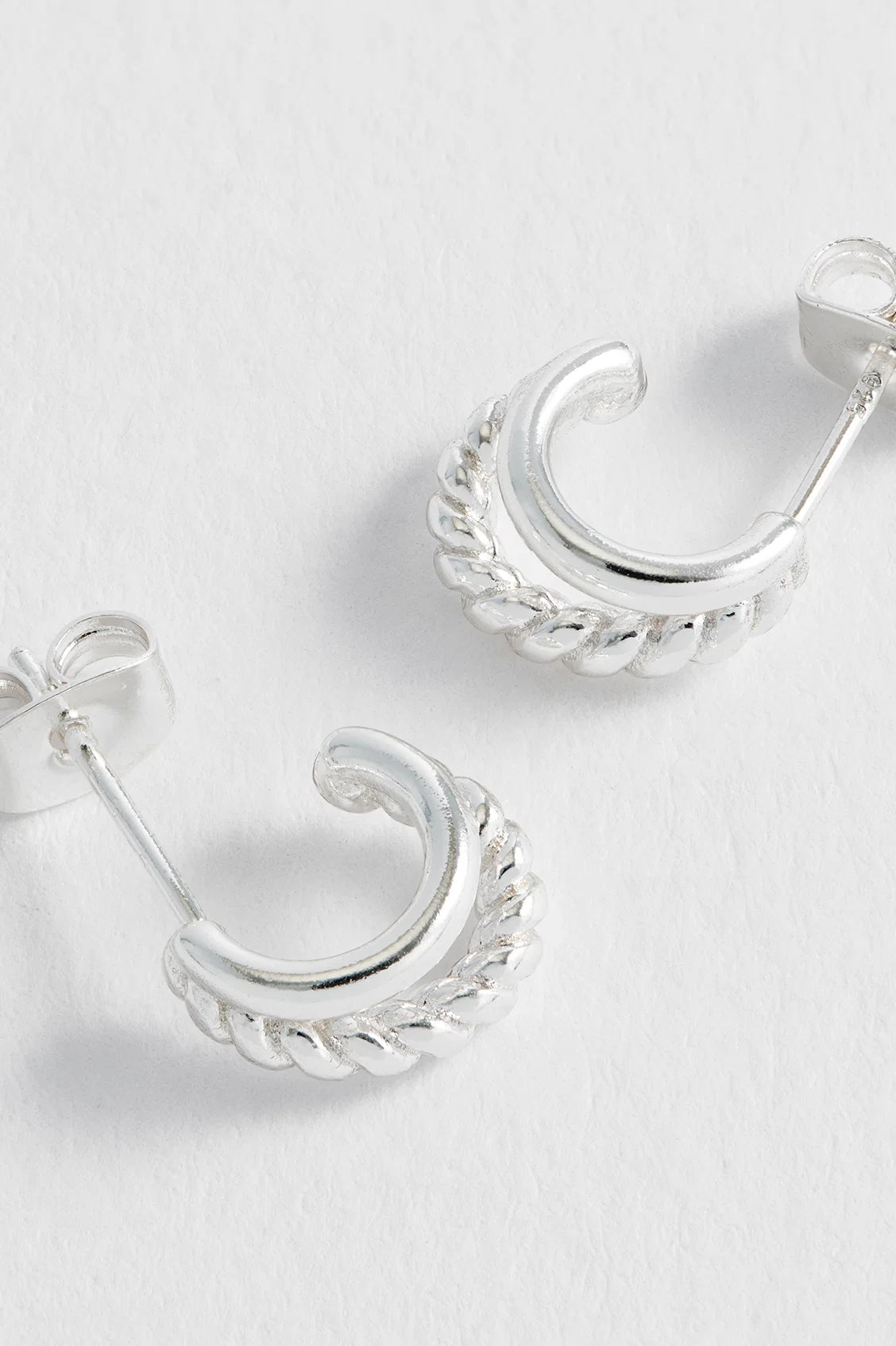 A pair of silver crescent-shaped hoop earrings with a twisted rope border detail, displayed on a textured white surface.