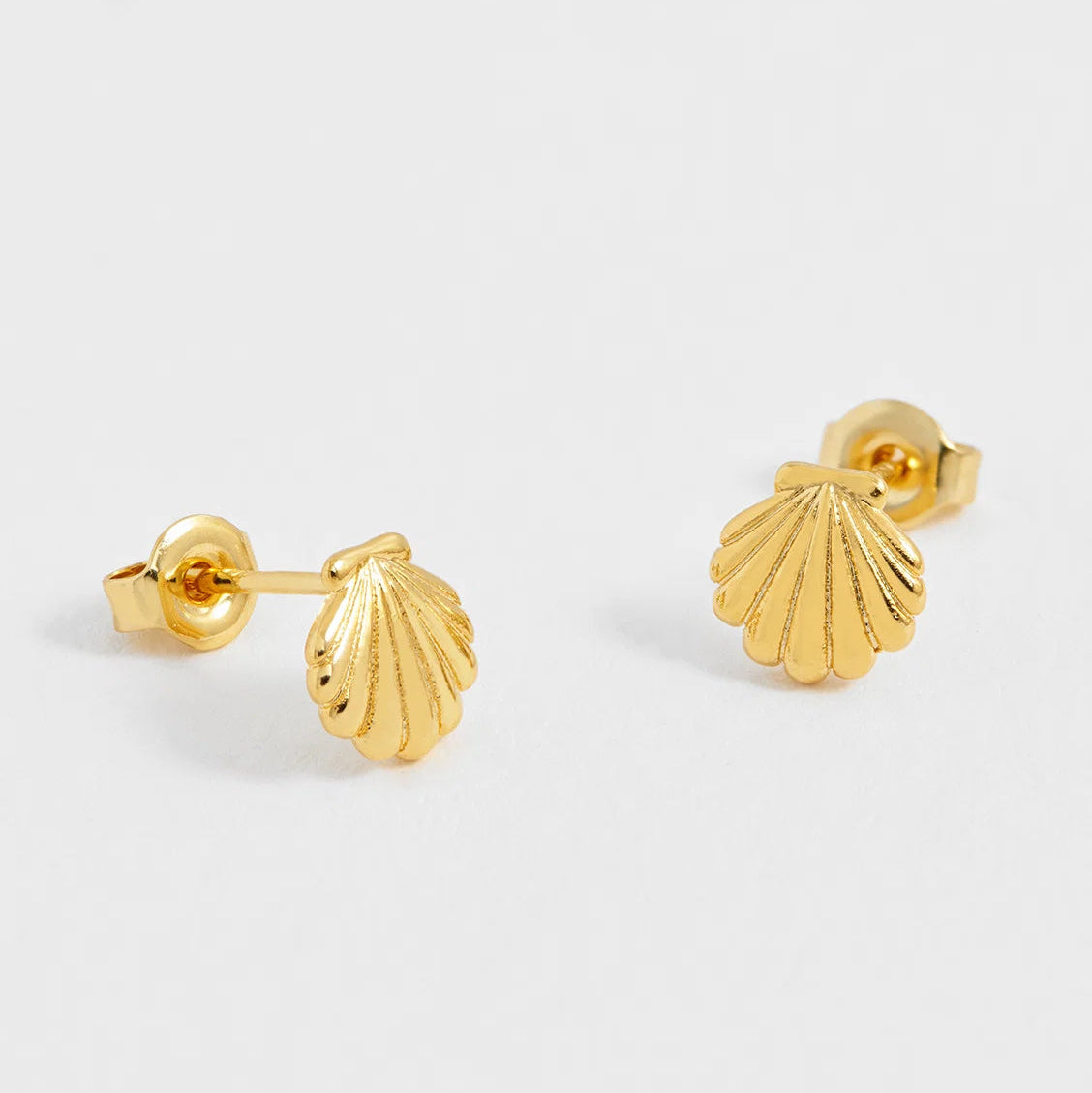 A pair of gold stud earrings shaped like ribbed scallop shells, shown on a clean light gray background.