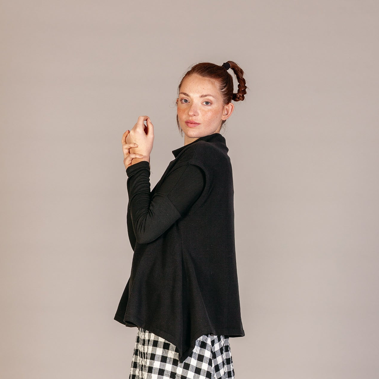 A woman modeling a black long-sleeve cardigan over a black and white gingham midi skirt, paired with striped socks and black patent leather shoes, against a solid light gray studio background.