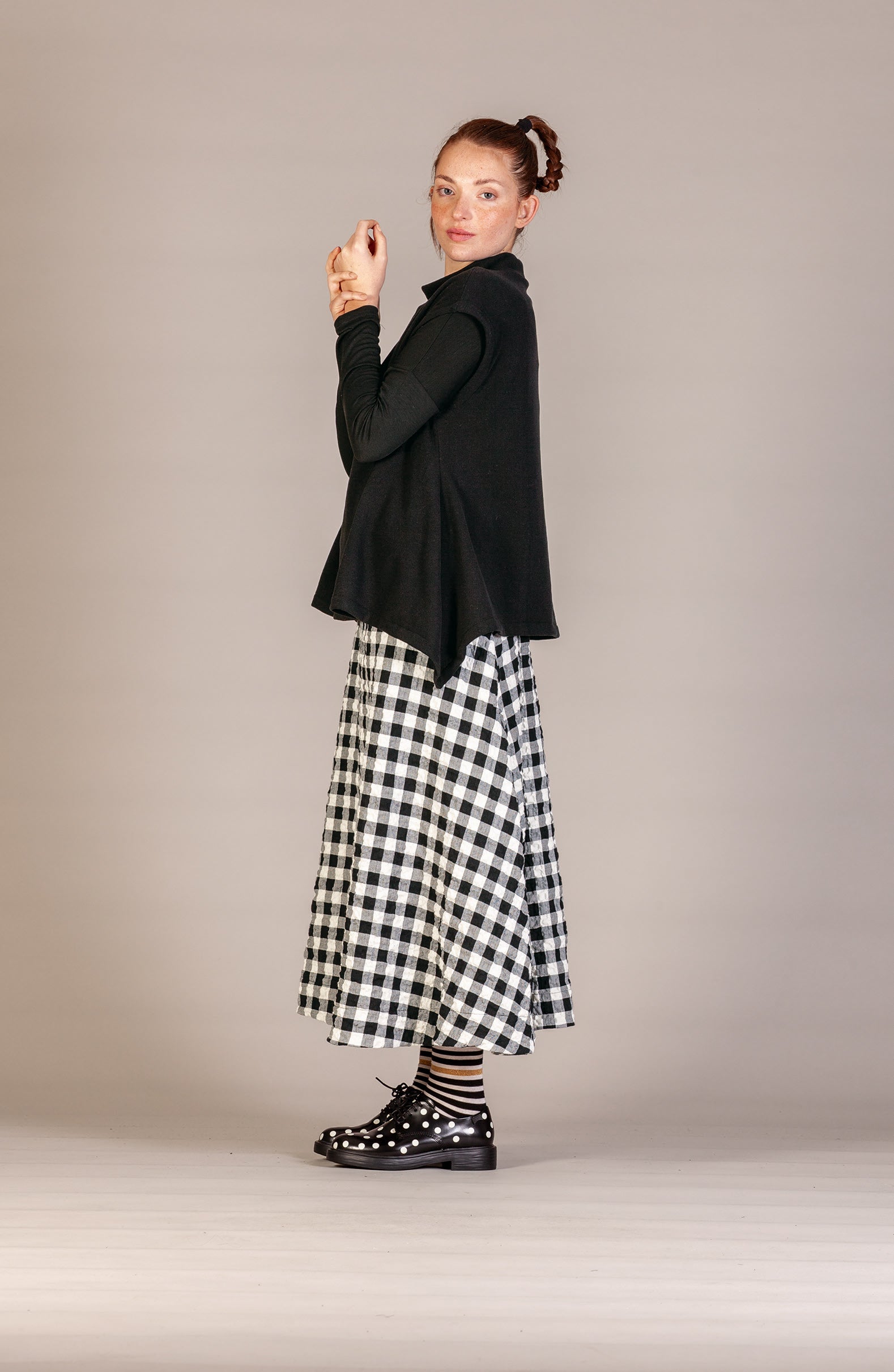 A woman modeling a black long-sleeve cardigan over a black and white gingham midi skirt, paired with striped socks and black patent leather shoes, against a solid light gray studio background.