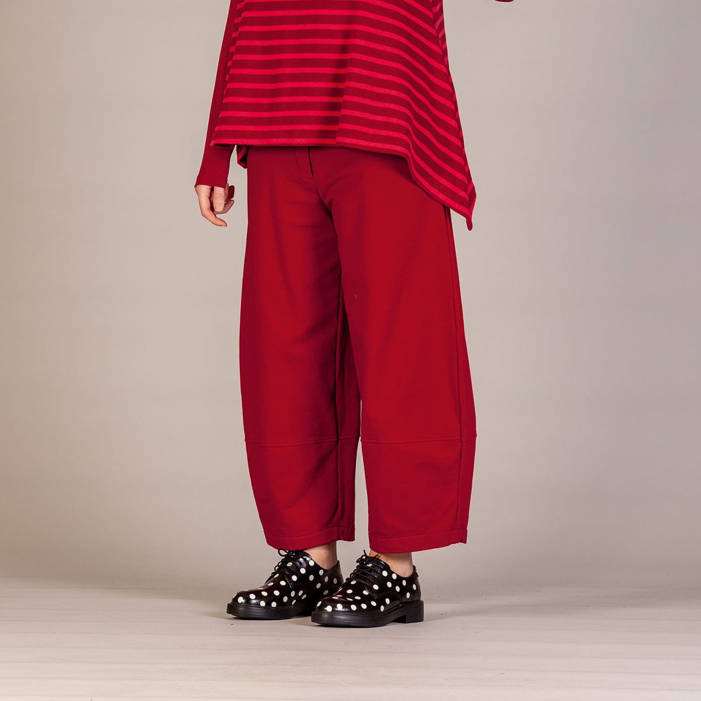 A model stands against a neutral studio backdrop wearing a red and dark red striped long-sleeve mock-neck top, matching wide-leg trousers, and black studded shoes.