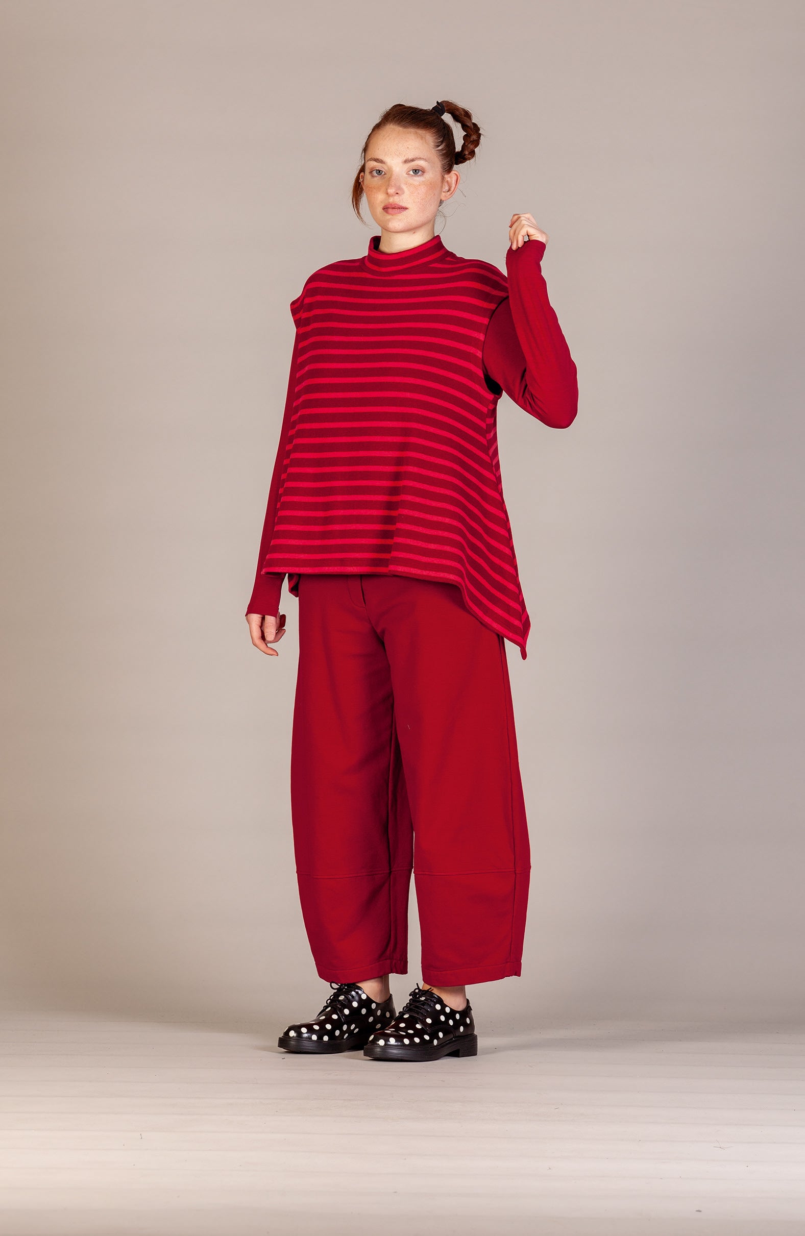 A model stands against a neutral studio backdrop wearing a red and dark red striped long-sleeve mock-neck top, matching wide-leg trousers, and black studded shoes.