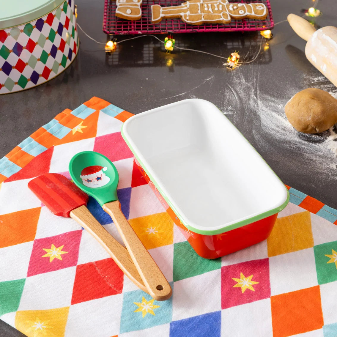 Colorful kitchen items including a spatula, spoon, and baking dish on a checkered tablecloth with Christmas decorations.