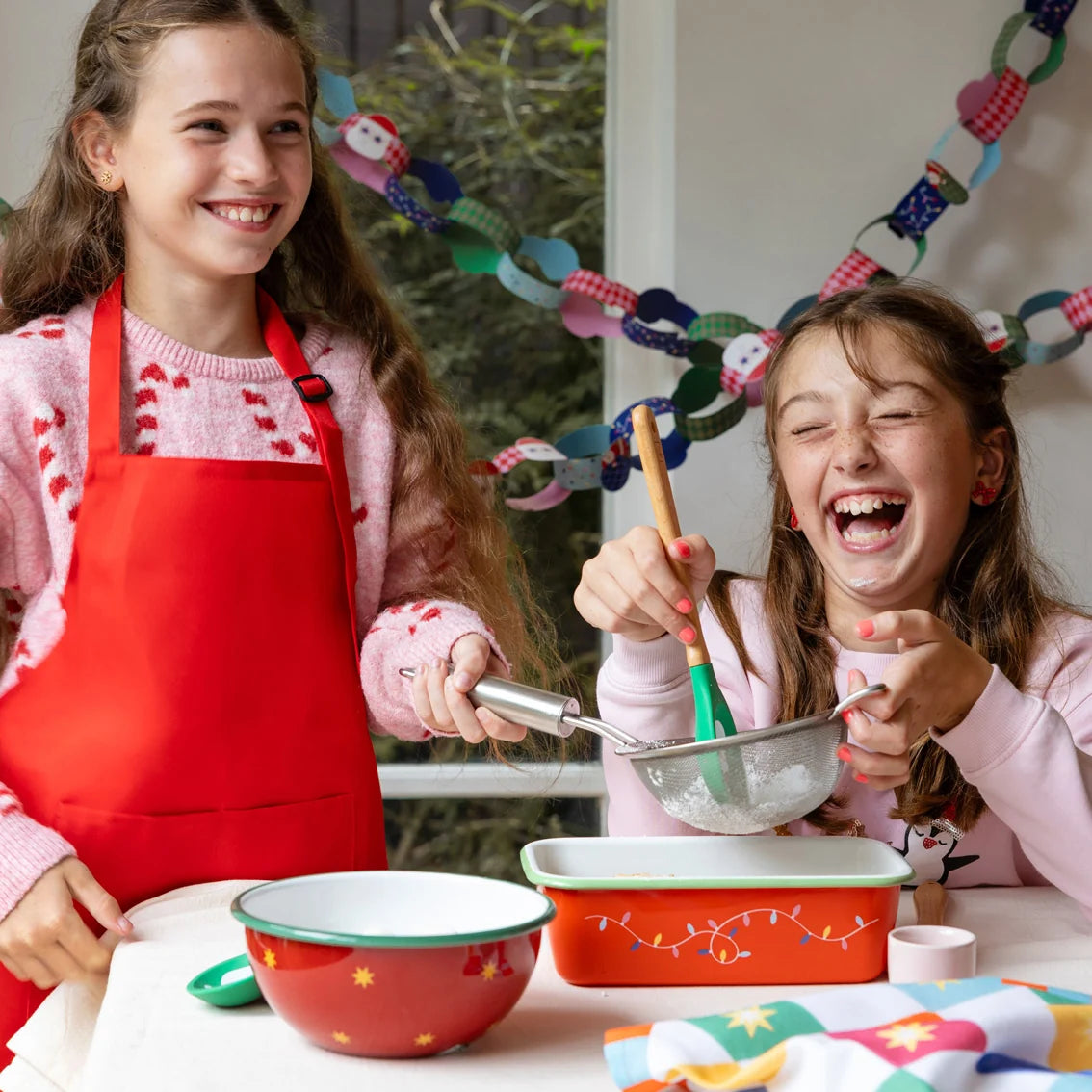 Two young girls smiling and laughing while baking together in a bright kitchen setting. One girl wears a red apron over a pink sweater, and the other wears a light pink top.