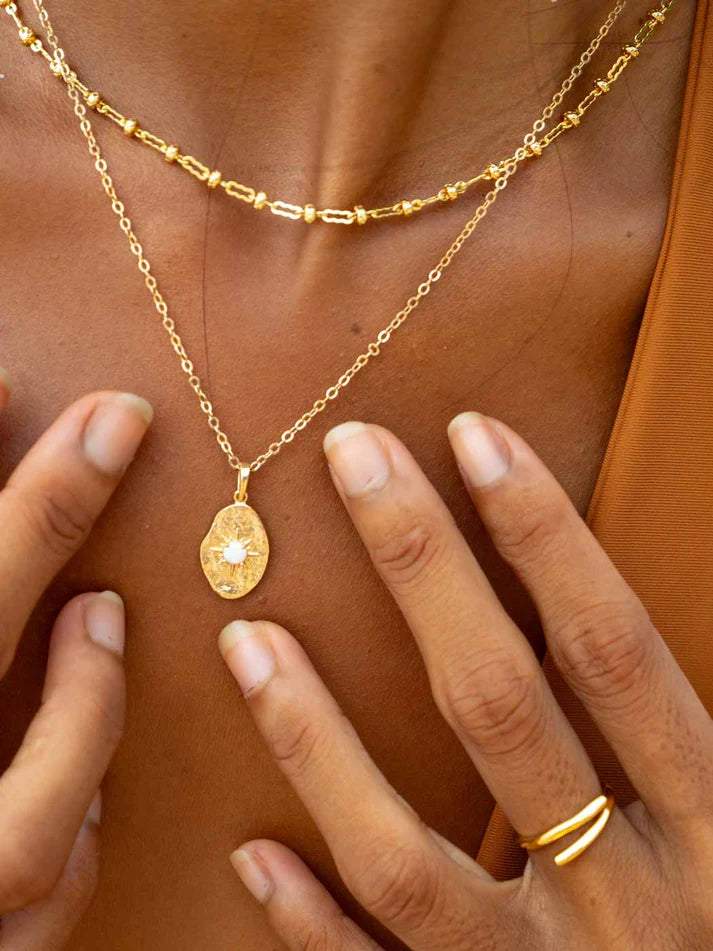 Gold necklace with a pendant and ring on a person's hand against a brown background