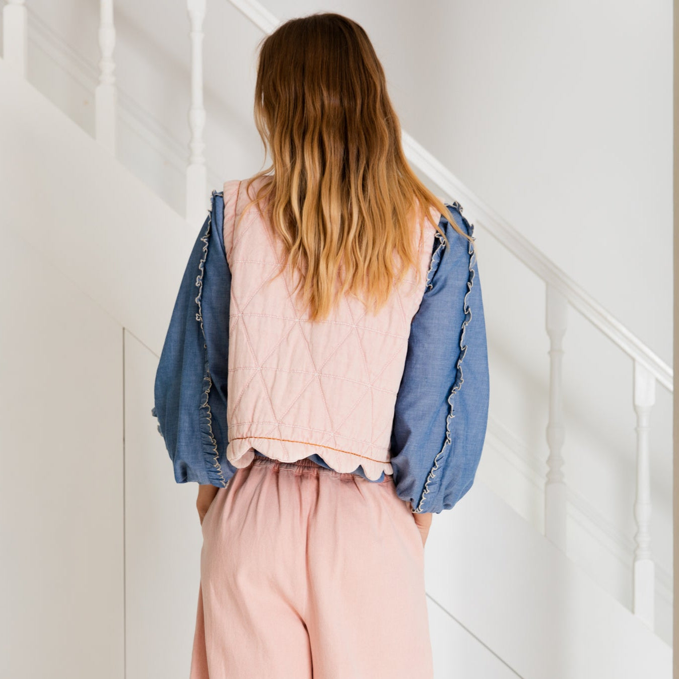 Pink quilted vest with scalloped hem, worn over a denim shirt and pink pants.