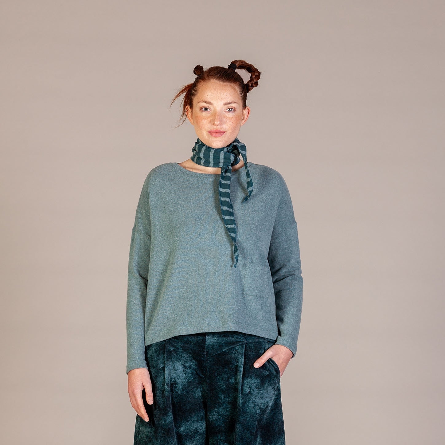 A model stands against a neutral beige studio background wearing a slate blue knit sweater, dark teal tie-dye wide-leg pants, a matching tie-dye scarf, and brown shoes.