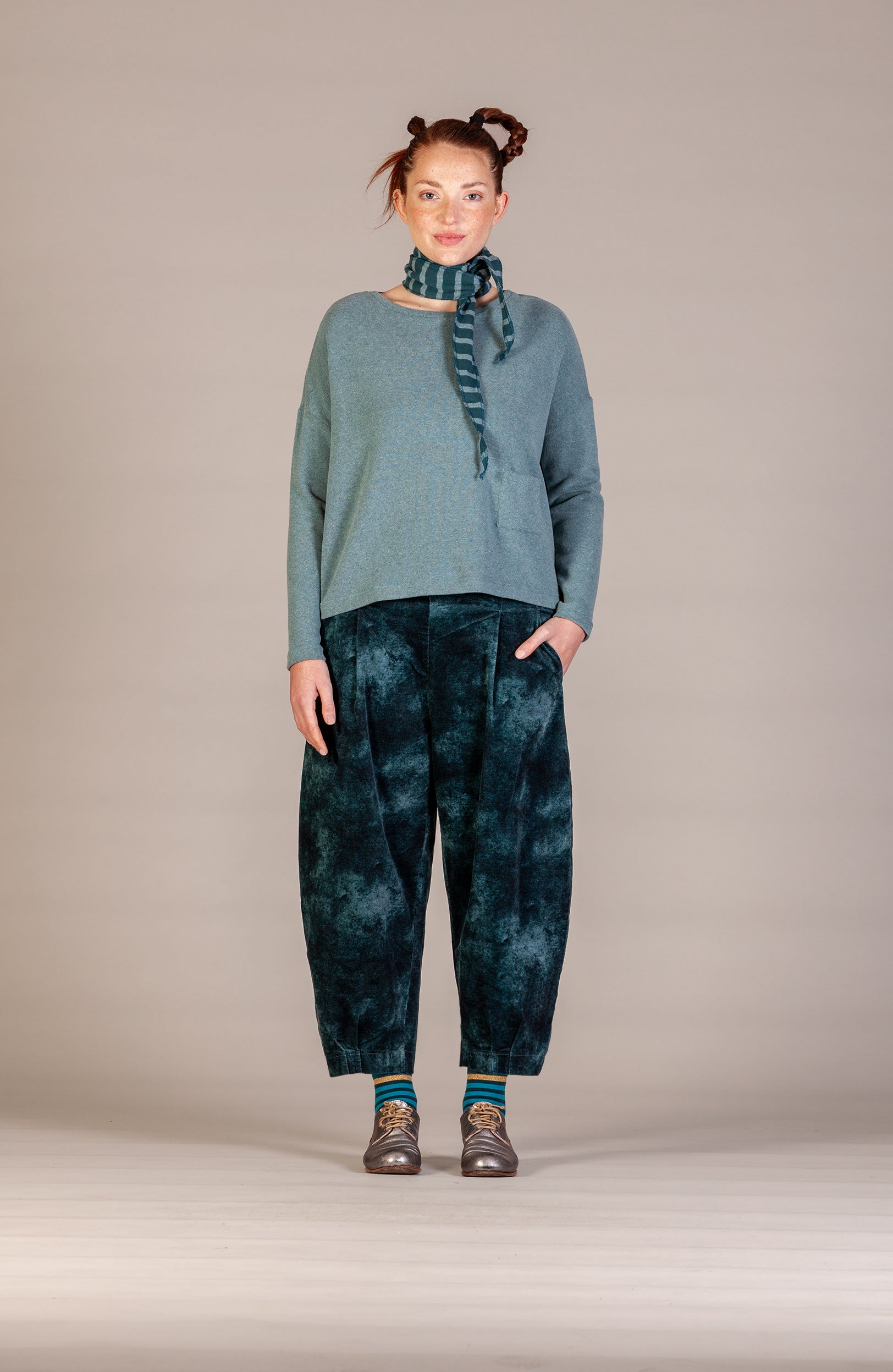 A model stands against a neutral beige studio background wearing a slate blue knit sweater, dark teal tie-dye wide-leg pants, a matching tie-dye scarf, and brown shoes.