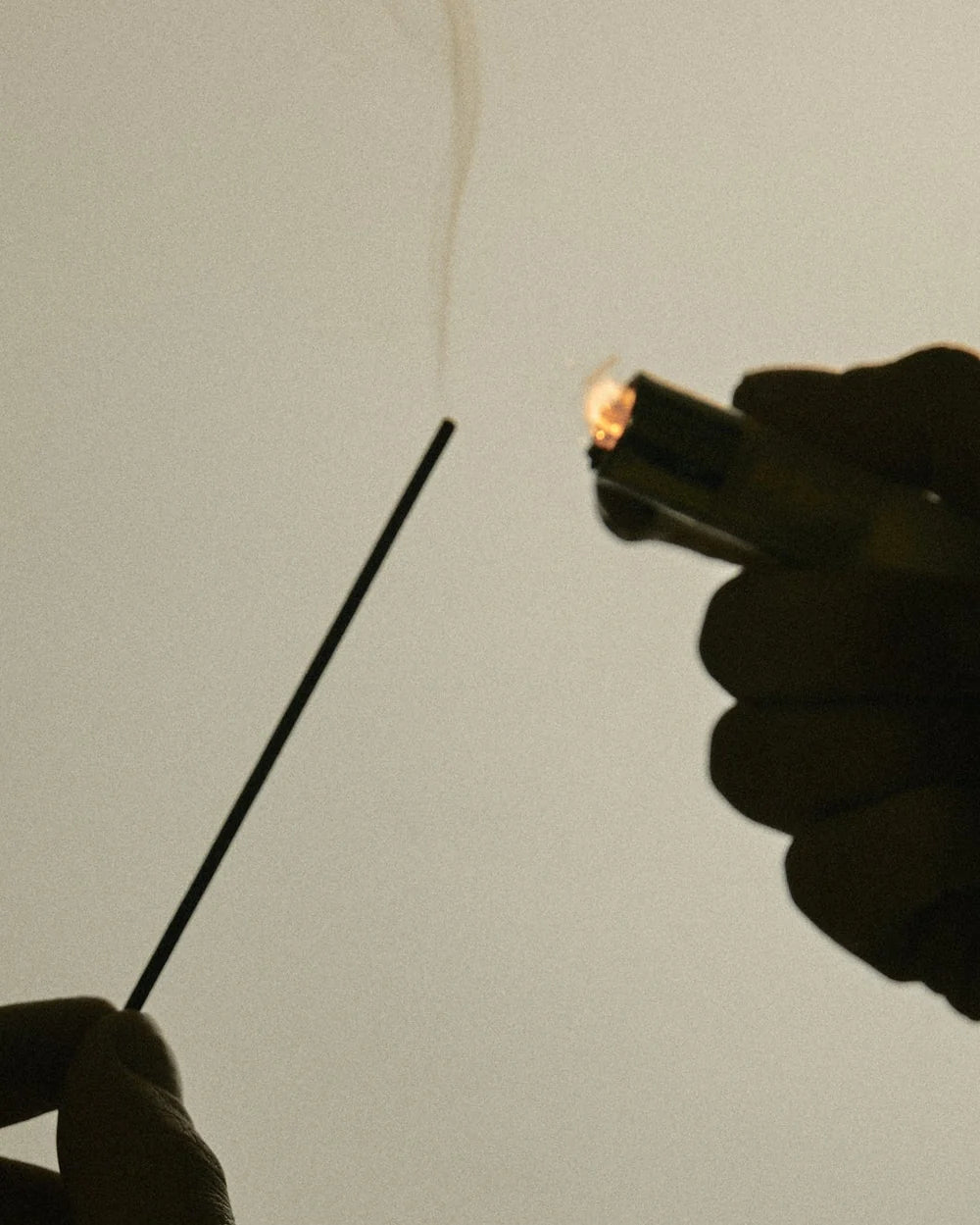 A close-up shot of a hand holding a lit incense stick with smoke rising, while another hand holds a small lighter against a plain light gray background.