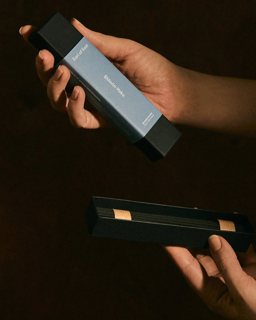 A hand holds a long rectangular box labeled 'Essence of Wood' and 'Quentin Volpi' against a dark brown background, with another open matching box below showing dark wooden sticks or strips inside.