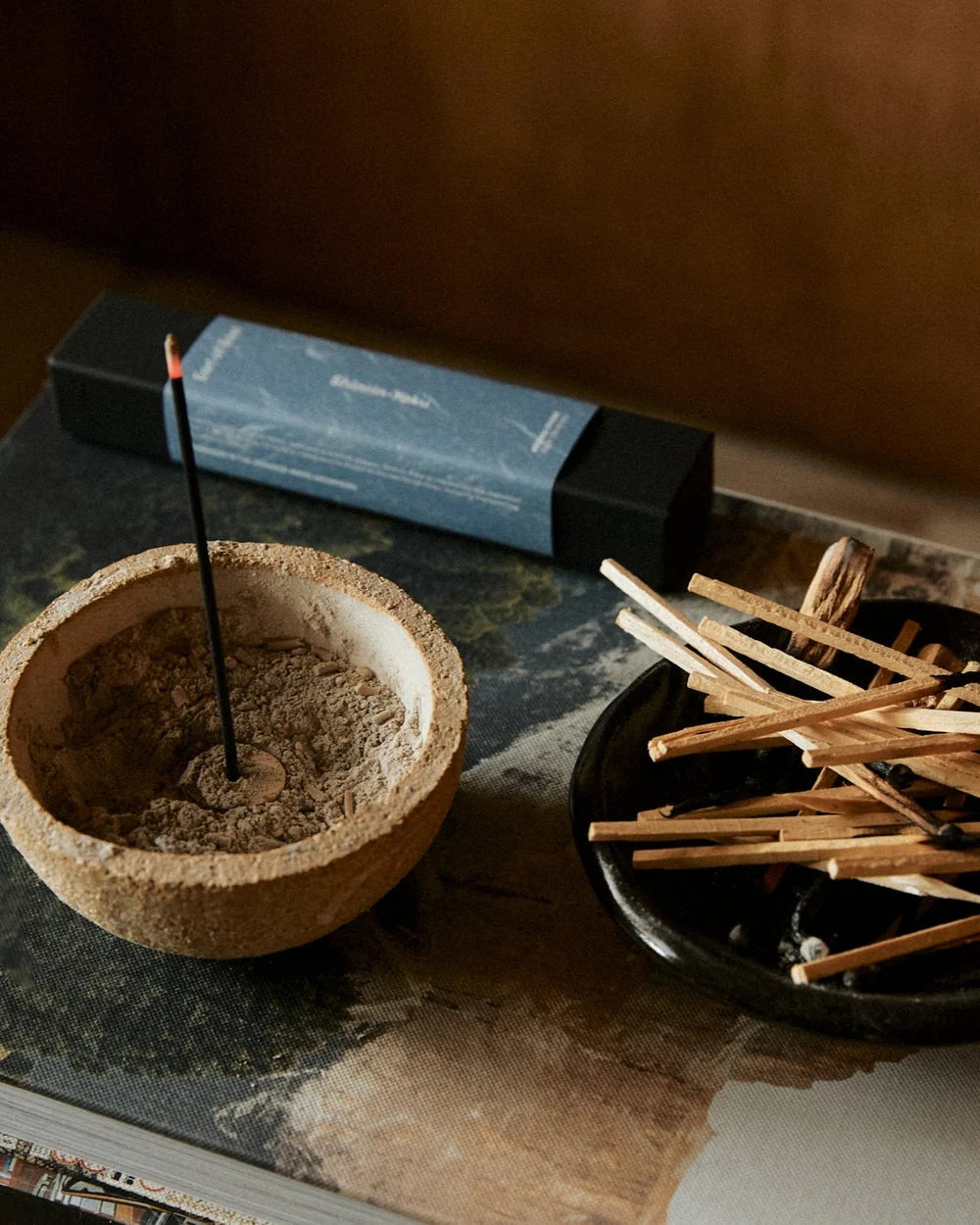 A close-up still life of incense and match accessories on a tabletop: a shallow round bowl filled with incense powder and a single incense stick standing upright, a black box with a blue label lying behind it, and a small black dish holding many wooden matchsticks.