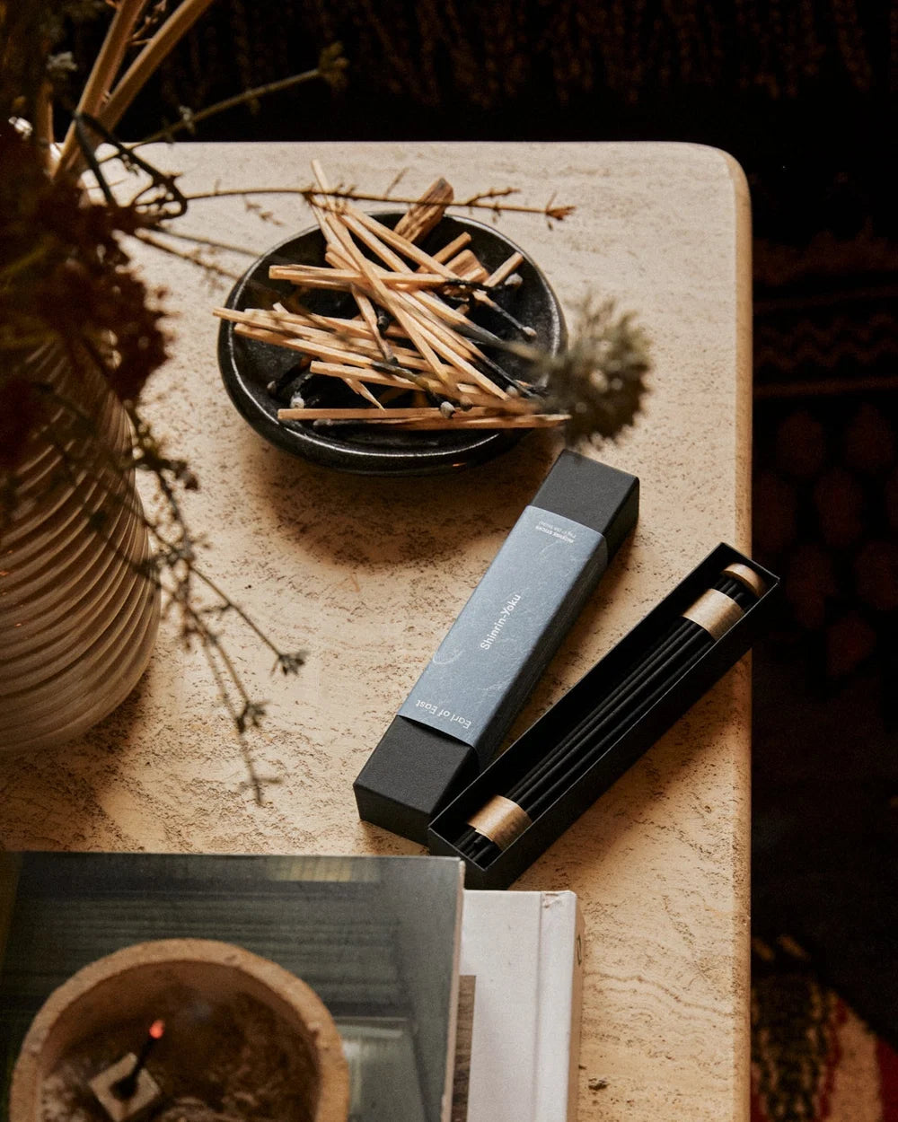A styled flat-lay of a black incense holder set on a textured beige stone surface, featuring a rectangular black box, an open black holder with incense sticks, a small black dish of dried grass and matches, and a ceramic vase with dried botanicals.