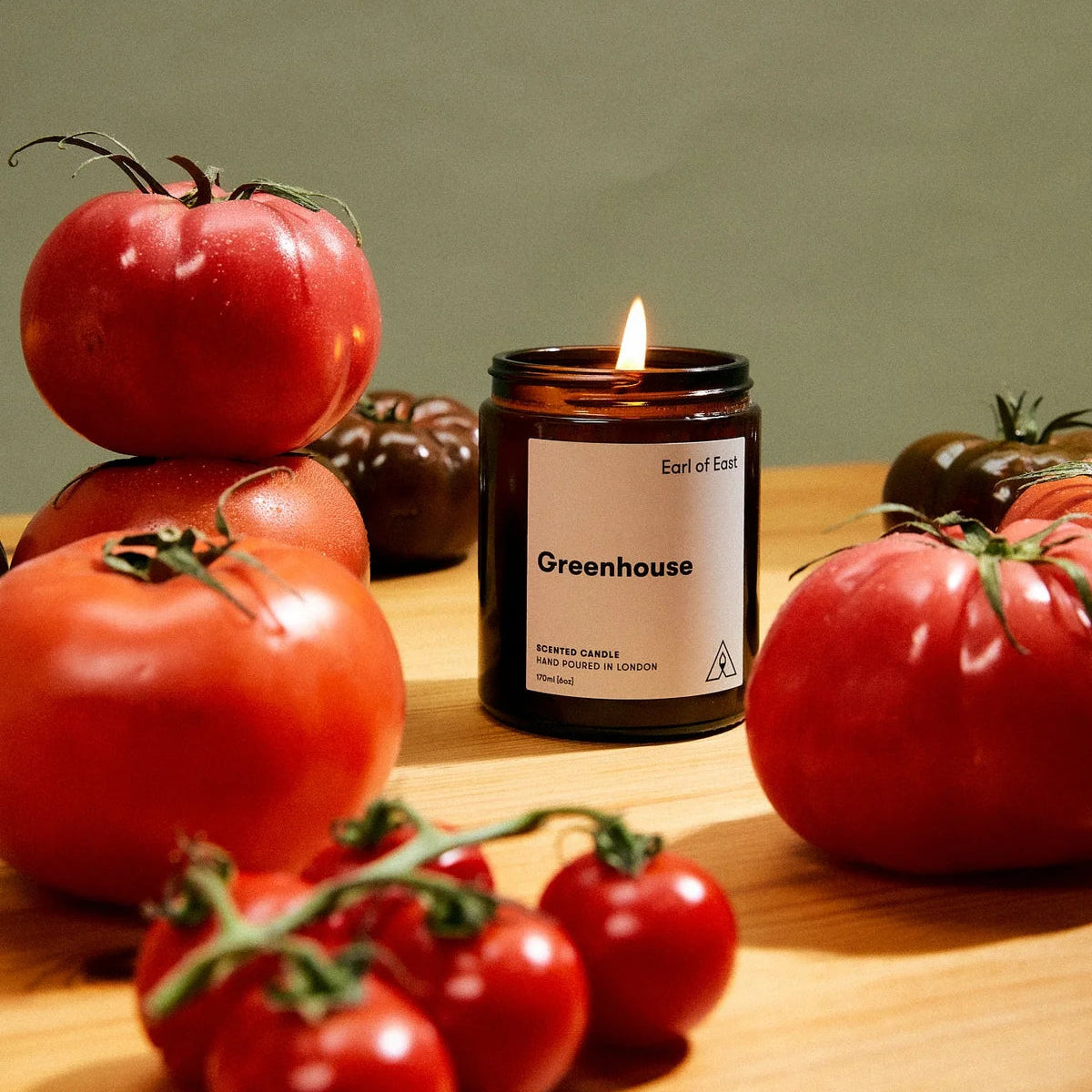 A lit amber glass jar candle labeled 'Greenhouse' by Earl of Sea, styled on a wooden surface with fresh red tomatoes around it.