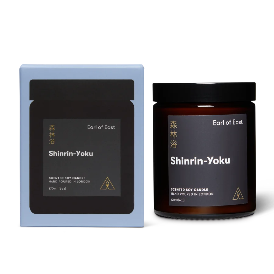 A scented soy candle by Earl of East in the Shinrin-Yoku scent, shown with its light blue rectangular box and an amber glass jar candle with a black lid on a white background.