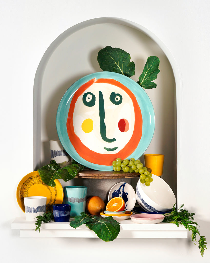 cream coloured plate with a painting of a face in deep blue, orange and green, set amongst other products from the FEAST range, designed by British-Israeli chef Yotam Ottolenghi, for Belgian design brand Serax.
