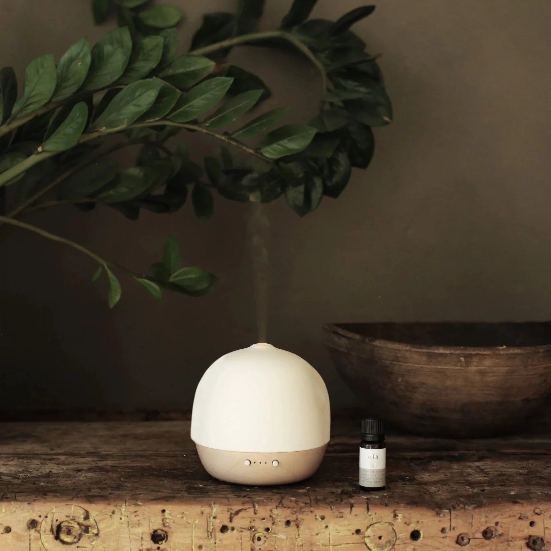 A beige and white ceramic essential oil diffuser emitting a gentle mist, next to a small bottle of essential oil.