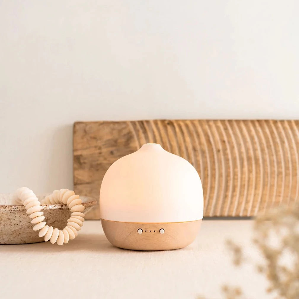 A modern, illuminated essential oil diffuser with a matte white top and light wood base.
