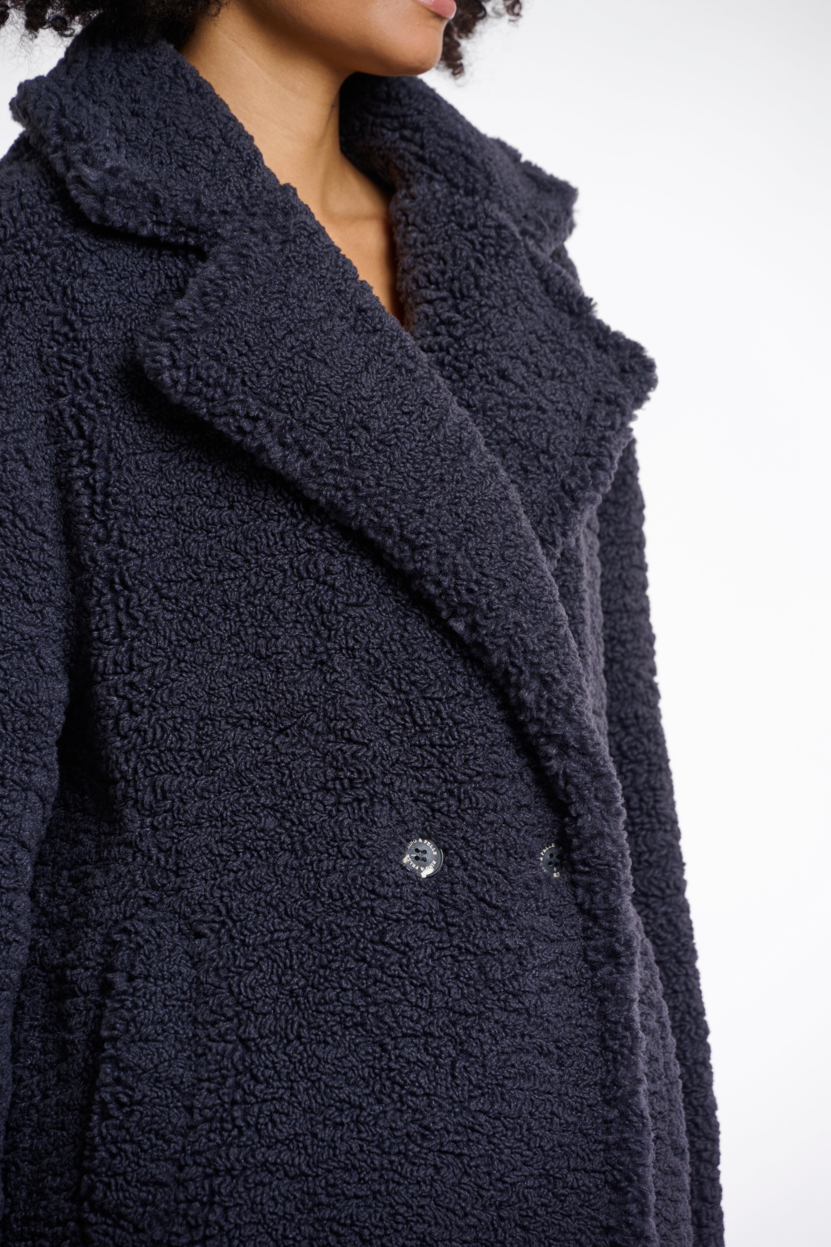 A woman models a double-breasted, teddy coat in night grey with a large lapel collar and side slit pockets.