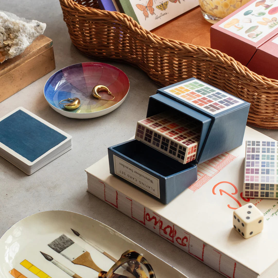 Color swatch book, dice, and small containers on a table with a wicker basket in the background.