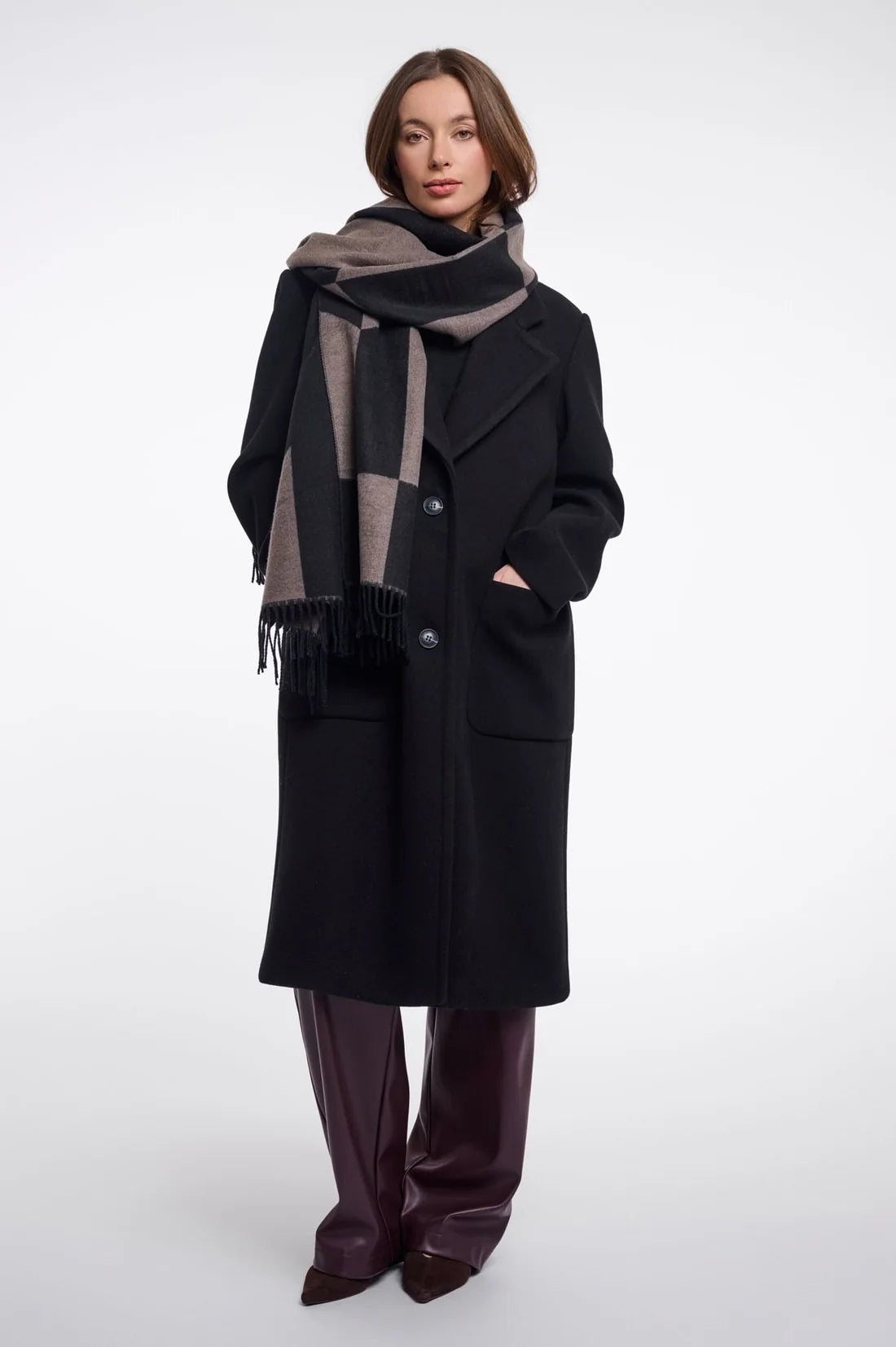 A woman modeling a long black wool coat with a large checkered scarf in black, gray, and taupe, paired with dark brown leather pants, against a plain white studio background.