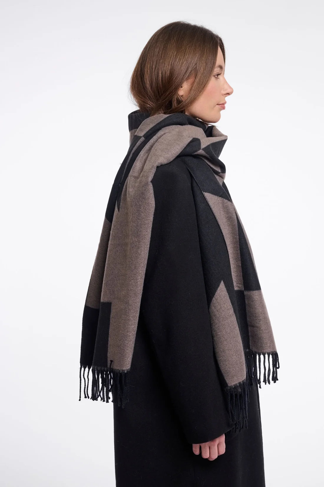 A woman shown in profile wearing a large woven scarf with a black and taupe-brown geometric block pattern and fringed ends, paired with a black coat against a plain light gray studio background.