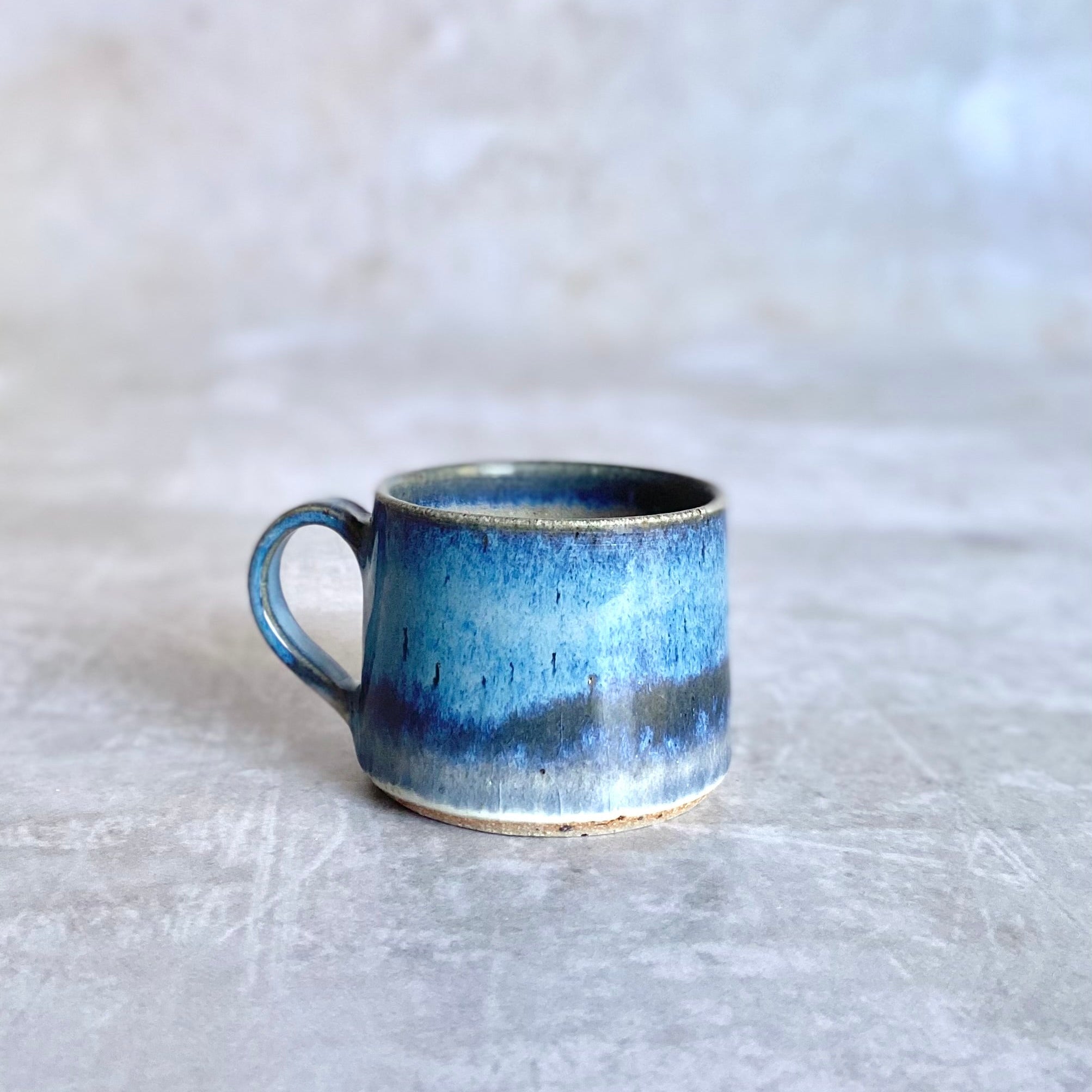 Small ceramic mug with a blue and grey glaze, featuring a textured, slightly irregular surface and a simple handle.