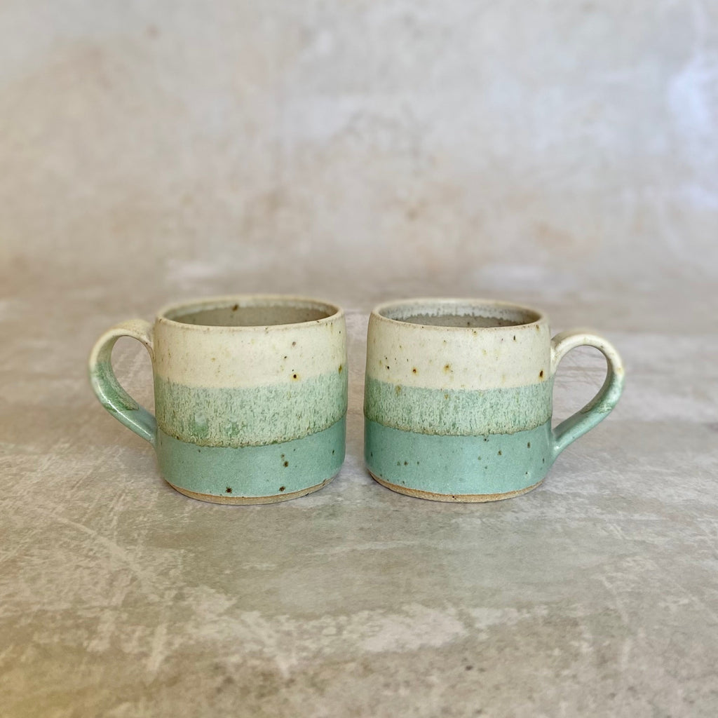 Two handmade ceramic mugs with speckled off-white, light green, and seafoam green bands.  Each mug has a sturdy handle.