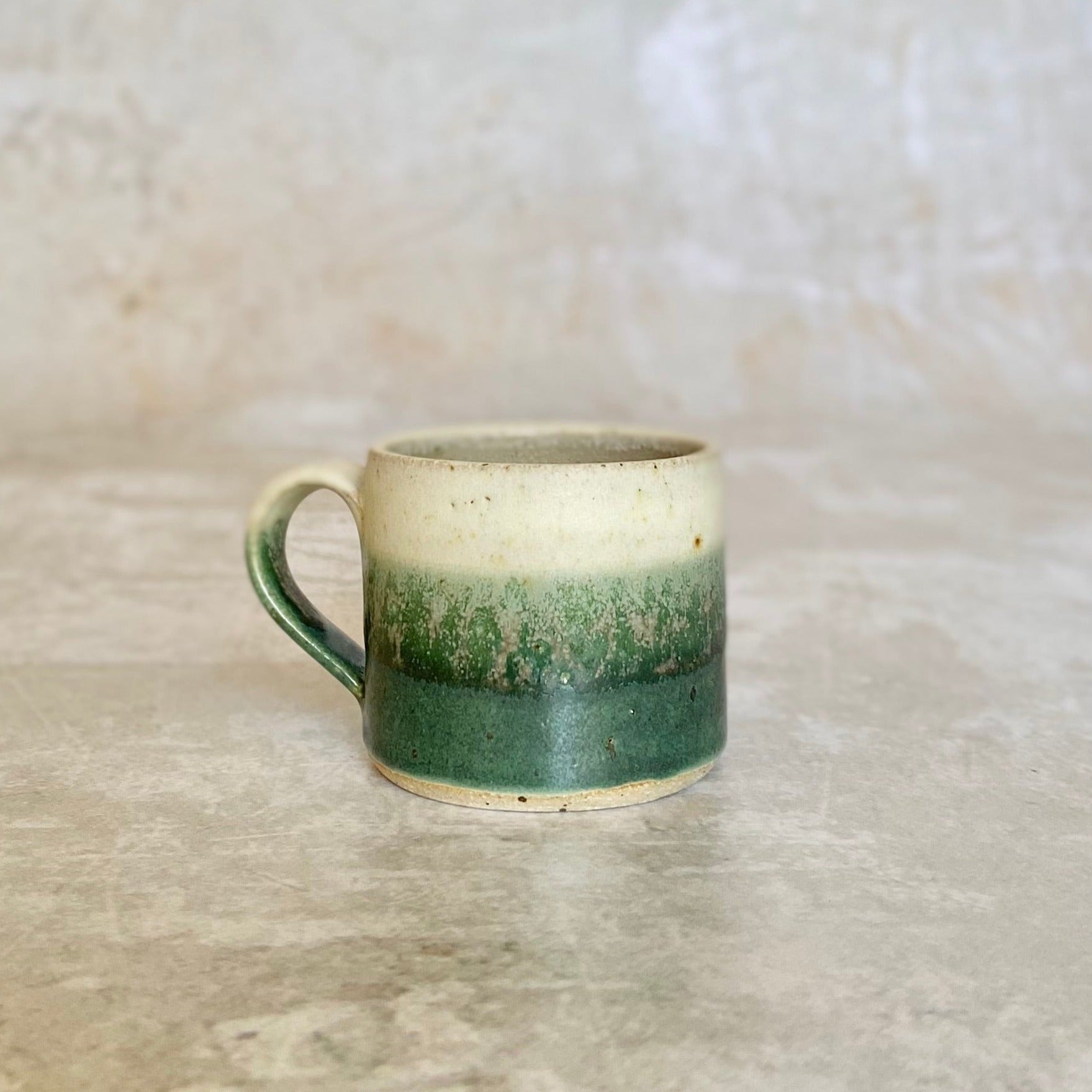 Small ceramic mug with a cream, green, and darker green ombre glaze.  The mug has a simple, slightly curved handle.