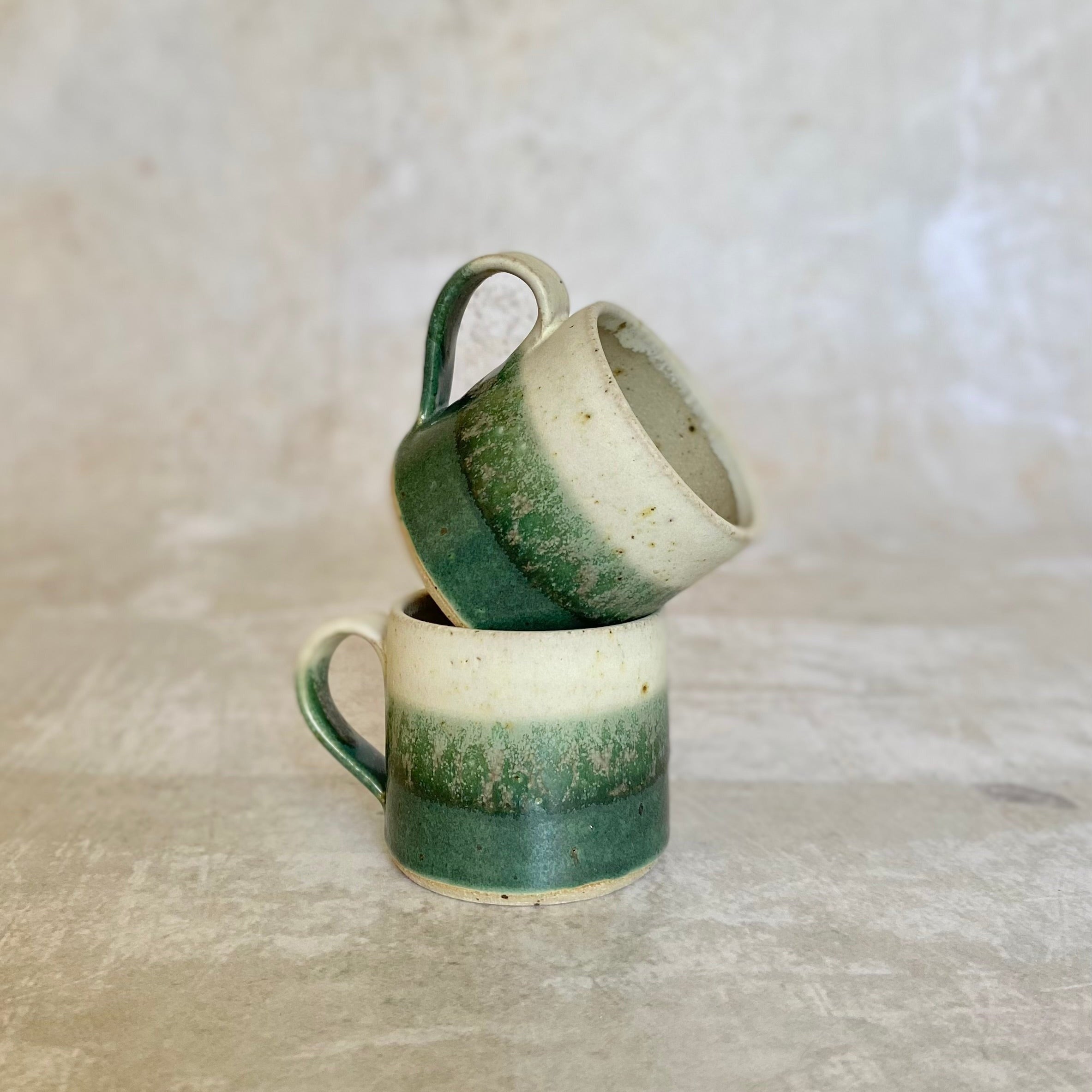 Two handmade ceramic mugs stacked;  bottom mug is green and off-white, top mug is similar but partially obscured.